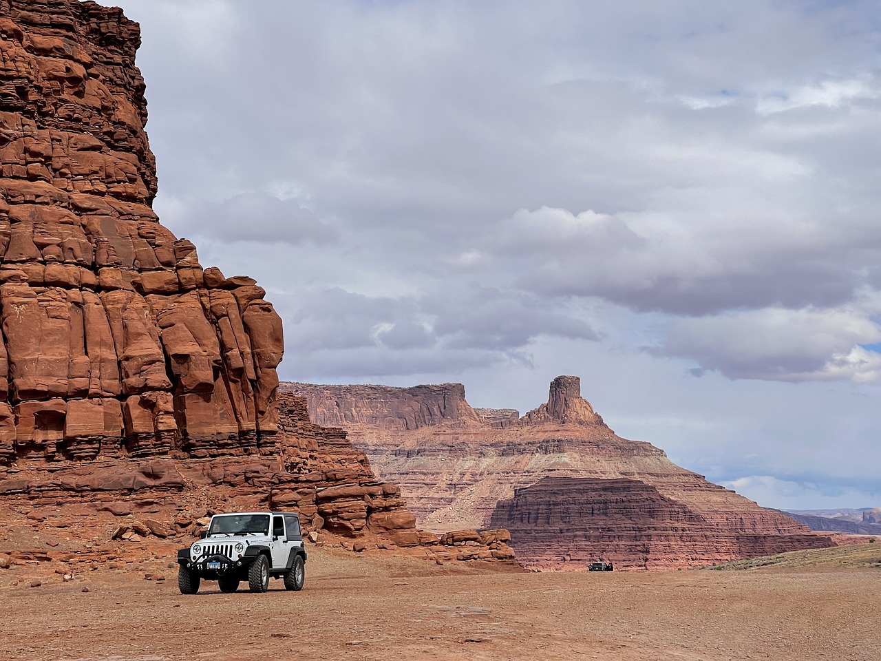 White Jeep parked on Jeep tour in a vast desert plain with towering red sandstone cliffs and buttes under a cloudy sky in Moab, Utah.